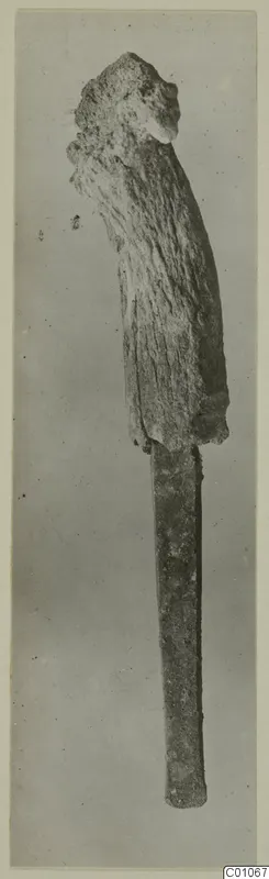 Bronze tool with a thick, crusted upper end and a long narrow flat blade, photographed upright against a grey background.