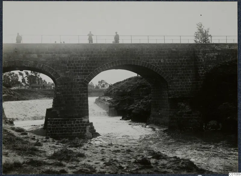 Brick bridge with two arches over fast-flowing water, with three people standing on its railing.