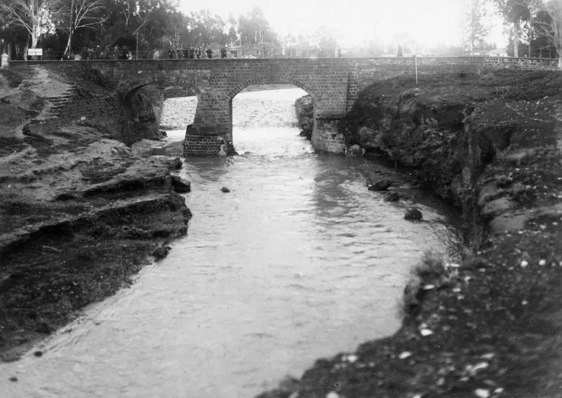 Fast-flowing river under a brick arch bridge at Nicosia, with onlookers standing along the parapet.