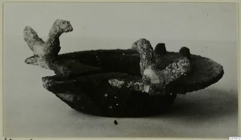 Corroded metal lamp with two upright projections, photographed from the side on a plain surface