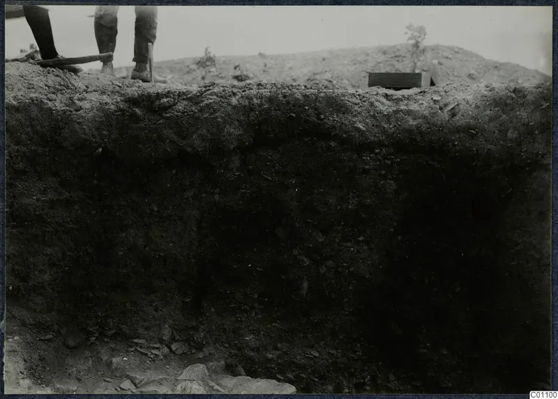 Vertical section of dark excavated soil at Idalion Acropolis, with the legs of two people and a tool handle at the top edge.