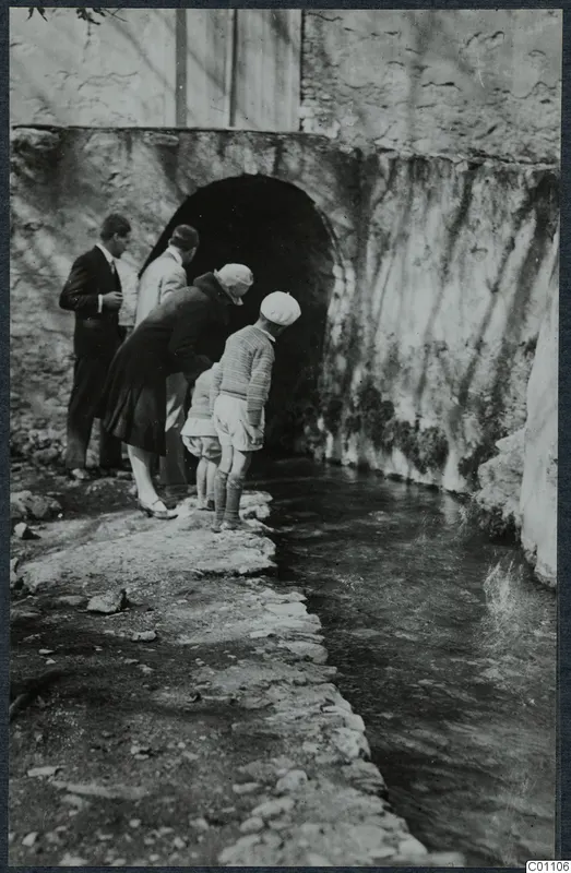 Visitors looking into a stream that flows from an arched opening in a stone wall at Kythrea
