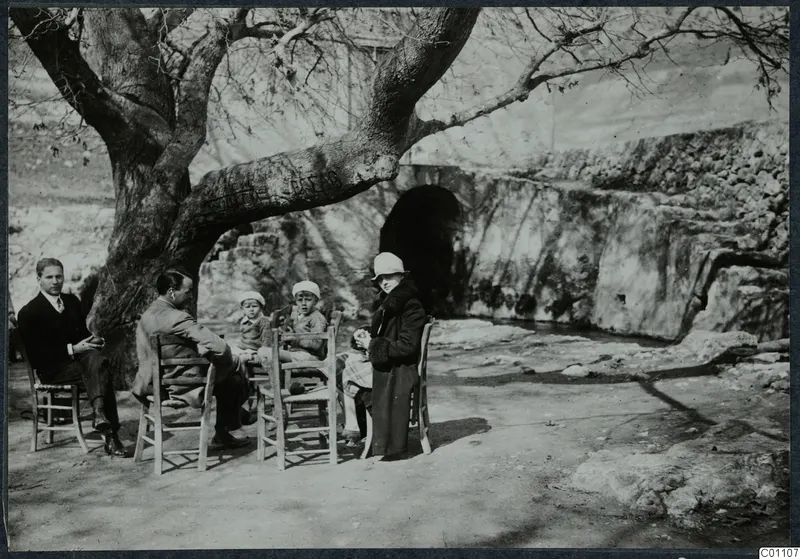 Group of people resting on wooden chairs under a bare tree beside a stone spring at Kythrea
