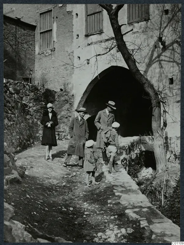 Two men, a woman and two children walk on a stony path past a vaulted archway in a stone building at Kythrea.
