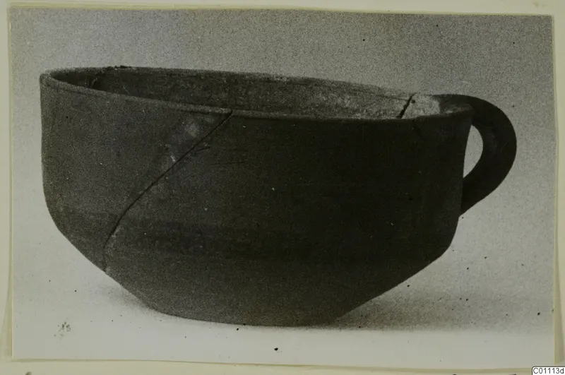 Pottery bowl with a single loop handle from the Kastros tombs at Lapithos, shown against a grey backdrop