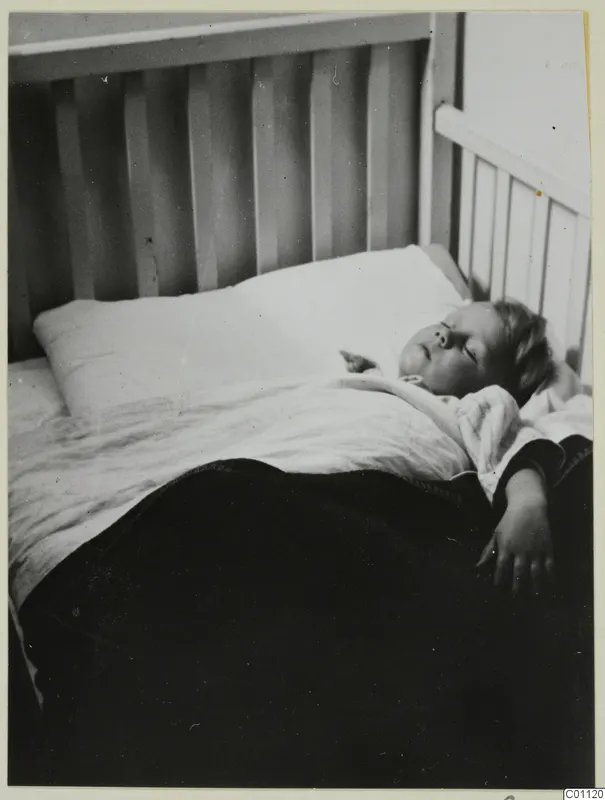 A young child asleep in a white-railed bed under a white pillow and dark blanket.