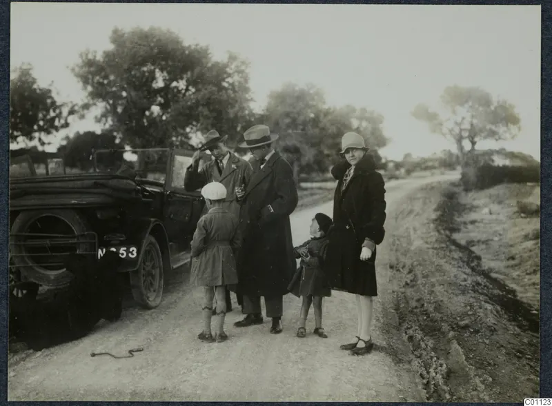 Two men, two children and a woman standing beside an open car on a dirt road at Idalion.