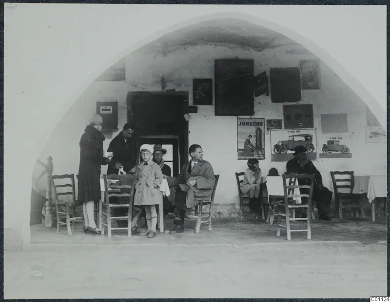Seated and standing people outside a whitewashed arched café at Skarinou, with posters and café chairs