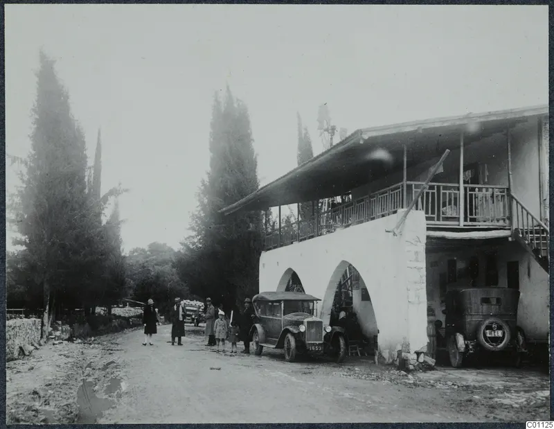 Group of people and two cars beside a two-storey white building with arches, on a road at Skarinou