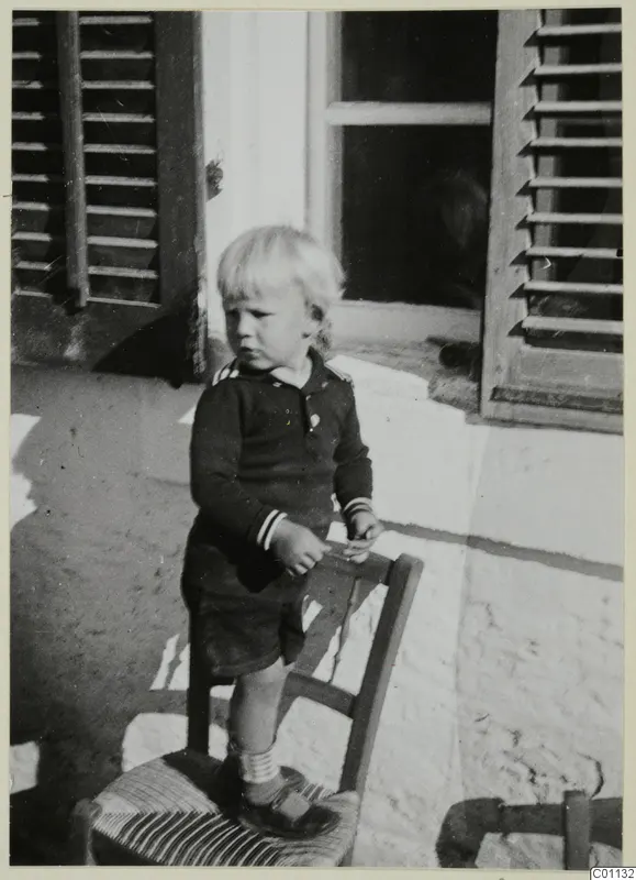 Small blond child in a dark sailor-style top standing on a wooden chair in front of a shuttered window.