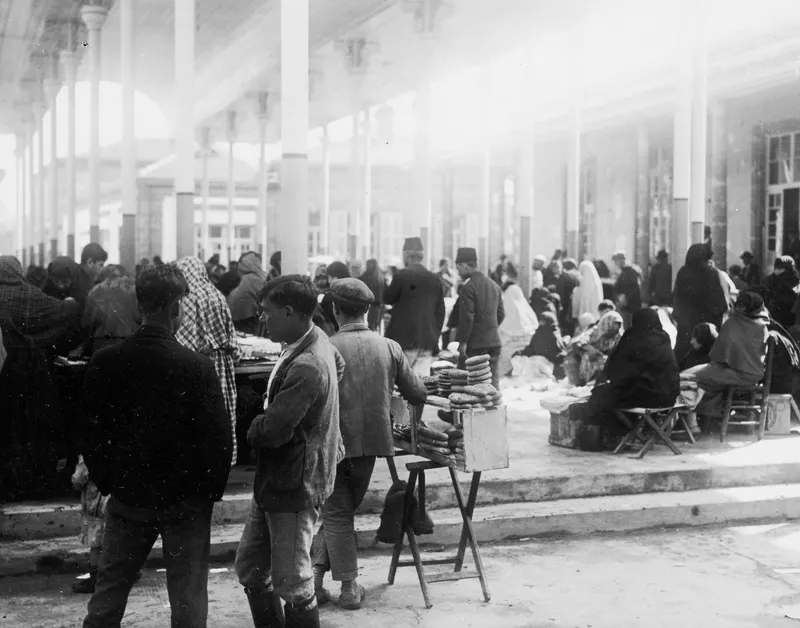 Market square in Nicosia with people in the shade of a colonnade, a vendor's stool stacked with bread rings in front.