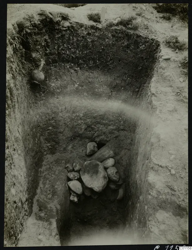 Tomb 5 at Marion during excavation, with a pile of stones and a vessel set in the trench wall.