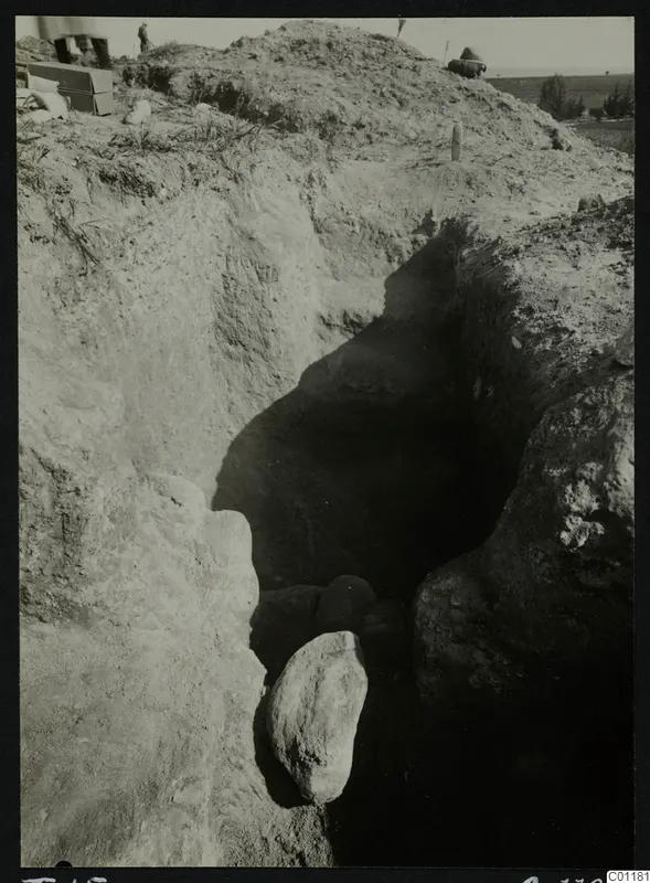 Excavation trench at Marion, Tomb 15, with a dark cut and large stones, people and grey boxes at the top edge.
