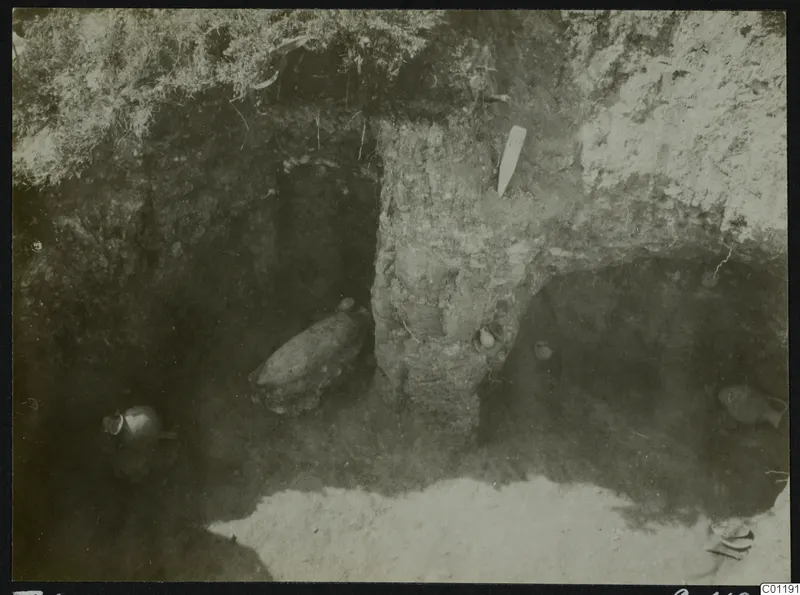 Excavated tomb at Marion with pottery vessels lying in situ on the chamber floor and a pale tag in the earth wall.