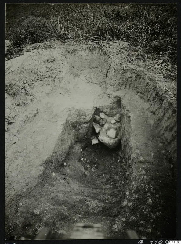 Excavated pit at Marion with a dromos-like cutting, grass above and a cluster of stones at the far end