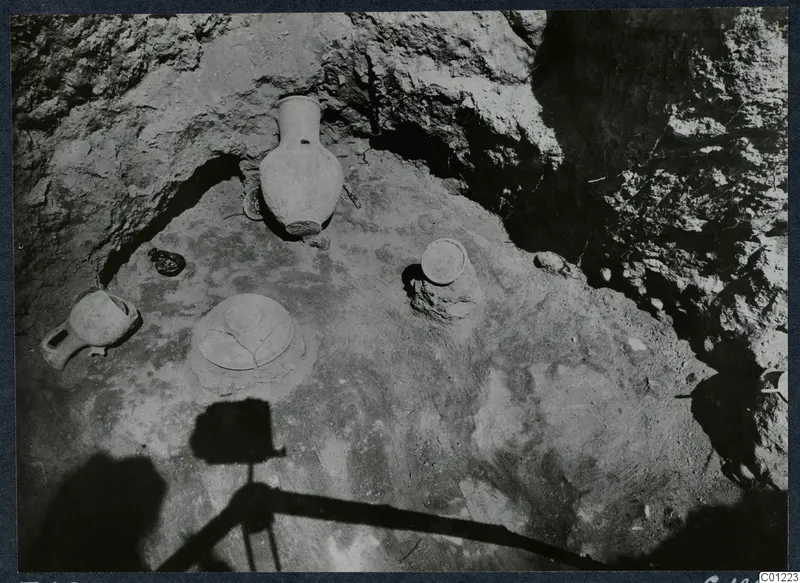 Tomb 45 at Marion during excavation, with pottery vessels and stones on the floor of a rock-cut chamber