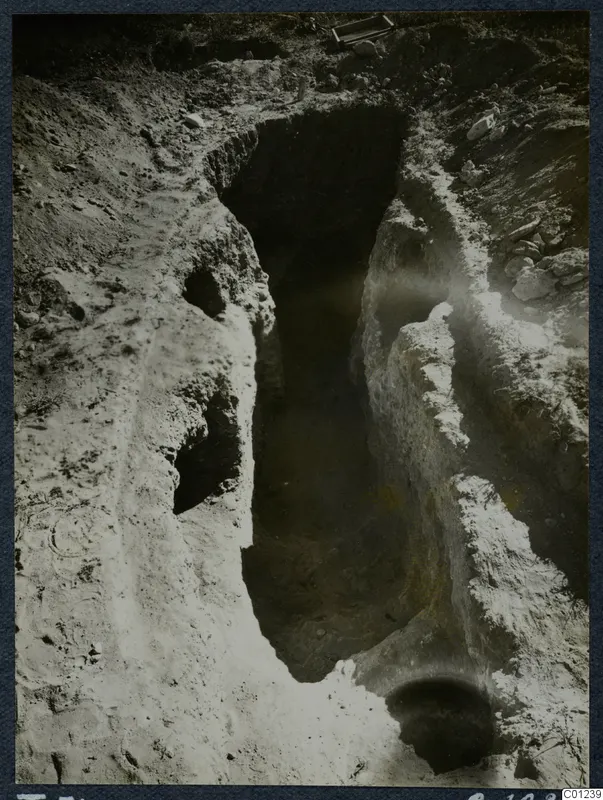 Excavation of Tomb 31 at Marion: a deep cut in pale soil with a dark opening in one wall and loose stones beside it.