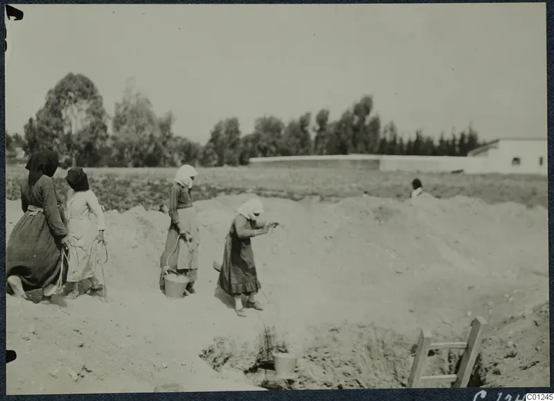 Women in headscarves and long skirts working in a sandy excavation trench at Marion, with a ladder at the edge.