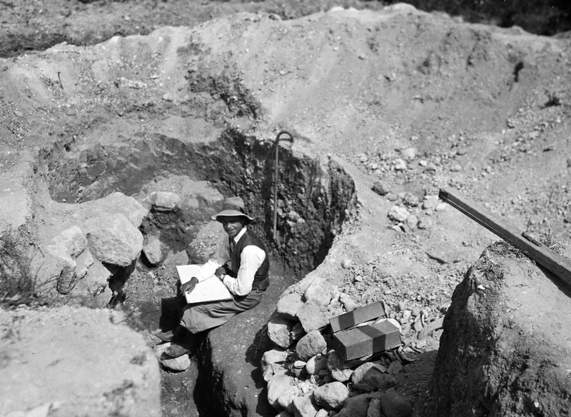 Erik Sjöqvist seated on the edge of an excavated cutting at Marion, resting a drawing board on his knees