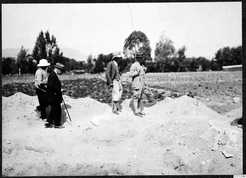 Four men stand and walk on a mound of excavated earth at Marion, with a cultivated field and trees behind.