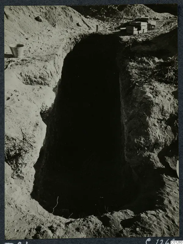 Tomb 21 at Marion during excavation, a deep rectangular cut with a bucket and stacked boxes at its edge