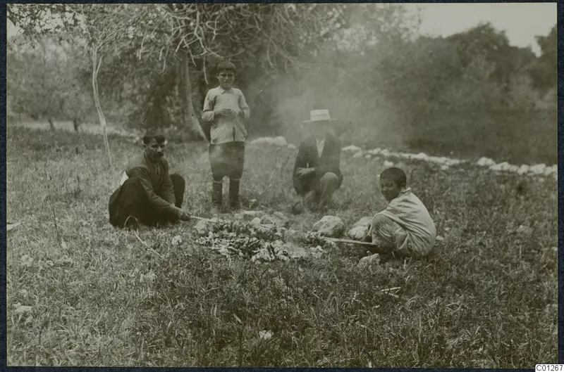 Four people at a small outdoor fire in a grassy field near Liso, with trees and a low line of stones behind them.