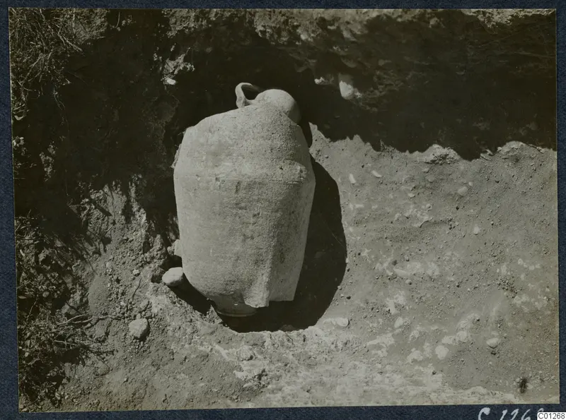 Large pale pottery vessel standing in an excavated cut at Marion, with stones around it