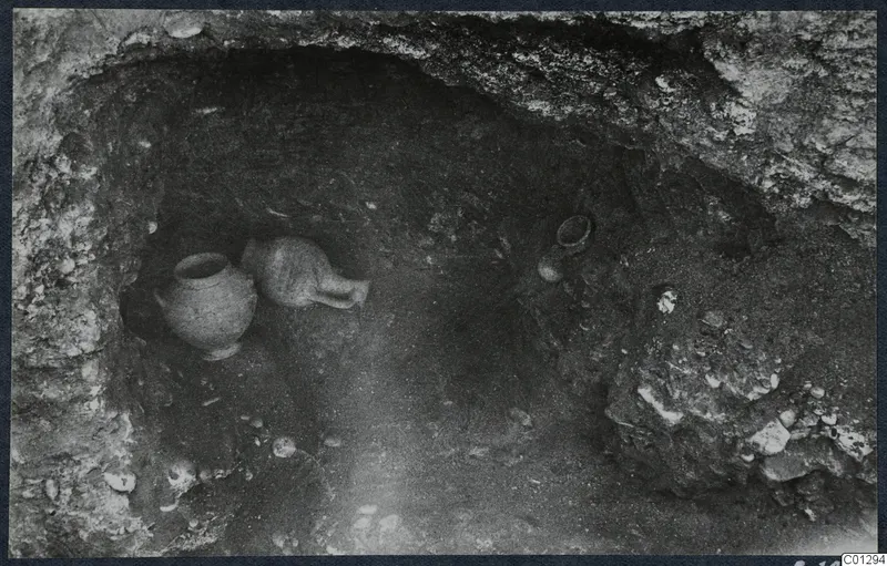 Interior of Tomb 59 at Marion during excavation, with two pottery vessels and loose stones on the floor of the cut.