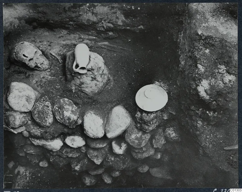 Tomb 59 at Marion during excavation, with a skull and pale vessels among stones and soil.