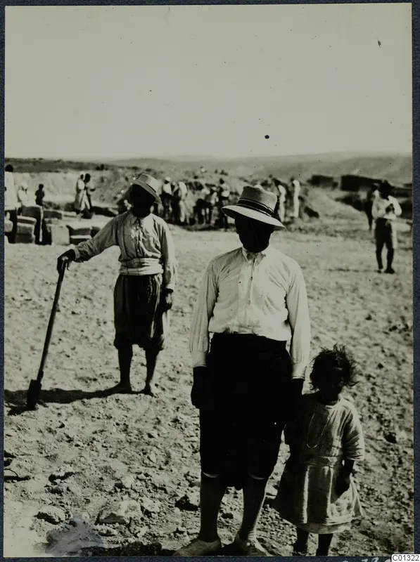 Two workers and a young girl on a dry excavation site at Marion, with more workers and stone blocks behind them.
