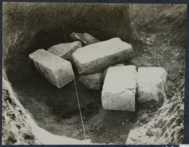 Excavated pit at Marion with several cut stone blocks lying in the soil, Tomb 77