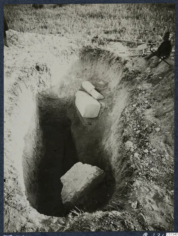 Tomb 74 at Marion during excavation: a deep rectangular cutting with stone blocks, and a seated person with tools at the edge