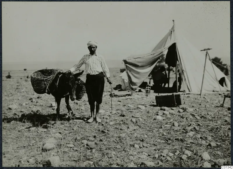 A man standing beside a donkey with wicker baskets, with a large white tent and a tripod at Marion
