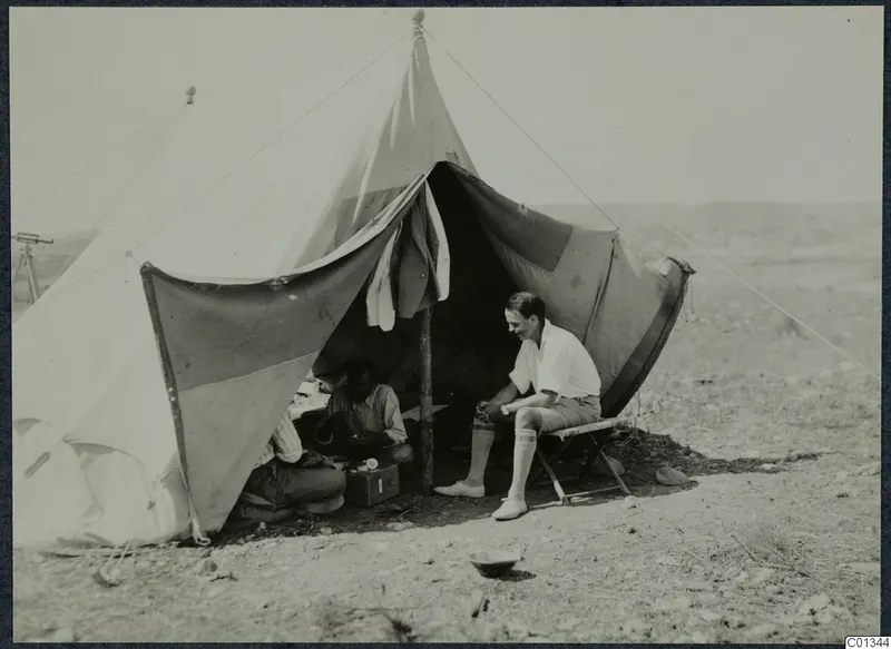 Man seated on a folding stool outside a large canvas tent at Marion, with two men working in the shade inside.