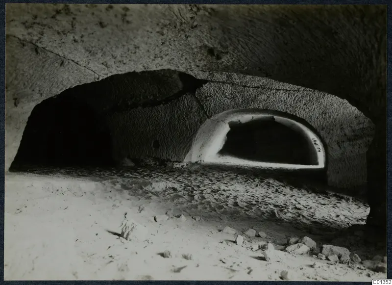 Interior of a rock-cut tomb chamber with an arched recess at the back and a sandy floor strewn with stones