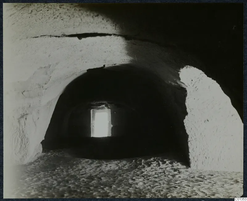 Interior of a vaulted rock-cut chamber, its floor strewn with sand and stones, with light from a doorway at the far end.