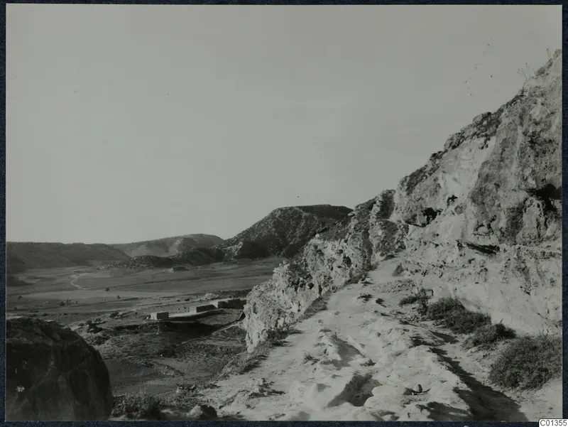 Dirt track running along a rocky cliff edge above a broad valley, with low stone walls on the valley floor