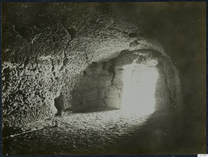 Interior of a rock-cut chamber with a bright opening at the back and a sandy floor, Karpas peninsula