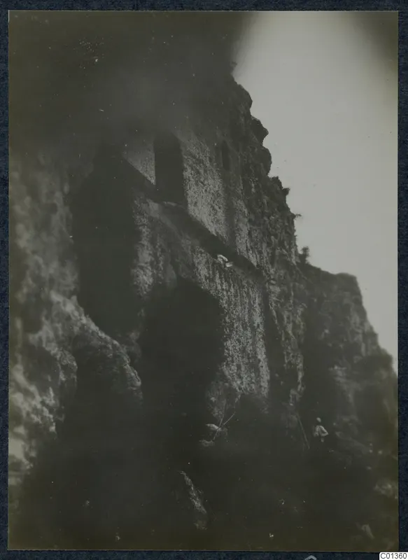 Dark view of a rocky cliff face with a square opening and two small figures on a ledge and at its foot