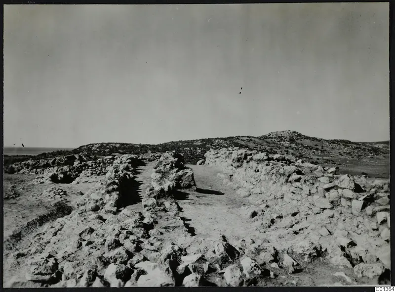 Rough stone walls and a dirt path running through a rocky excavation site toward low hills, with the sea at the far left.
