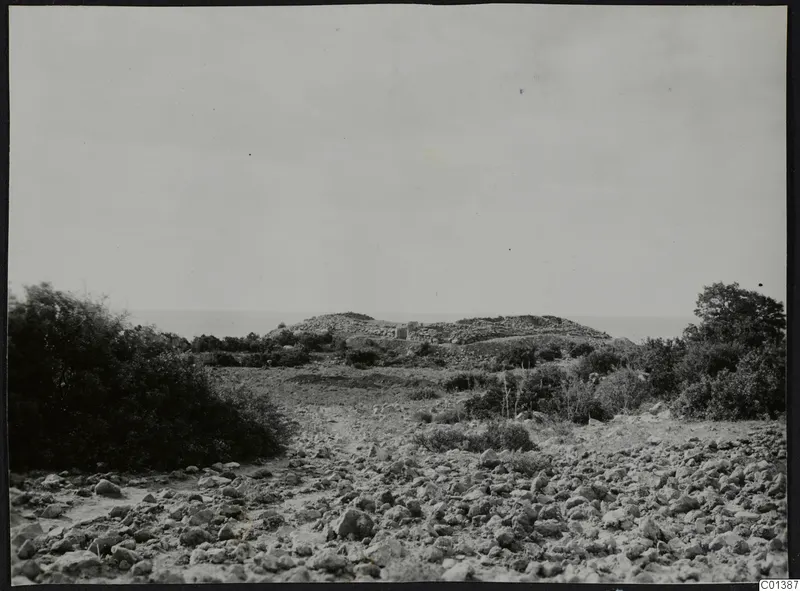 Rocky ground with scrub in the foreground and a low stony mound with a small structure against the sea at Nitovikla