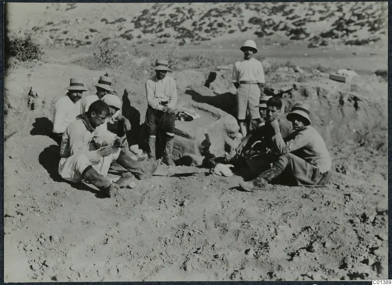 Several men in hats sit and stand on sandy ground beside a large stone block at Oura.