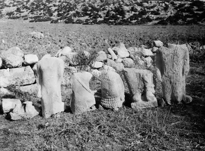 Several limestone statues and fragments standing in grass against a stone wall at Oura, with a field behind.