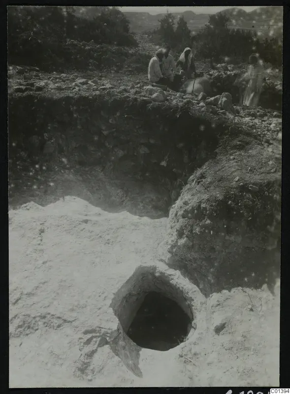 Excavation at Nitovikla on the Karpas Peninsula, with a round opening in pale soil and people seated among stones above