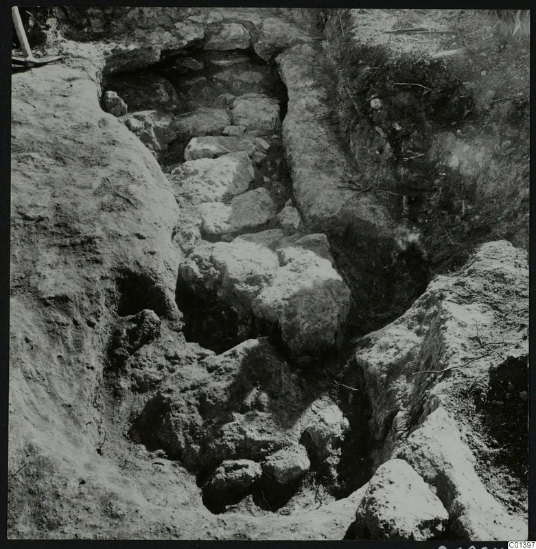 Excavation at Nitovikla on the Karpas Peninsula, with large stones in a trench and a pick in the top left corner.