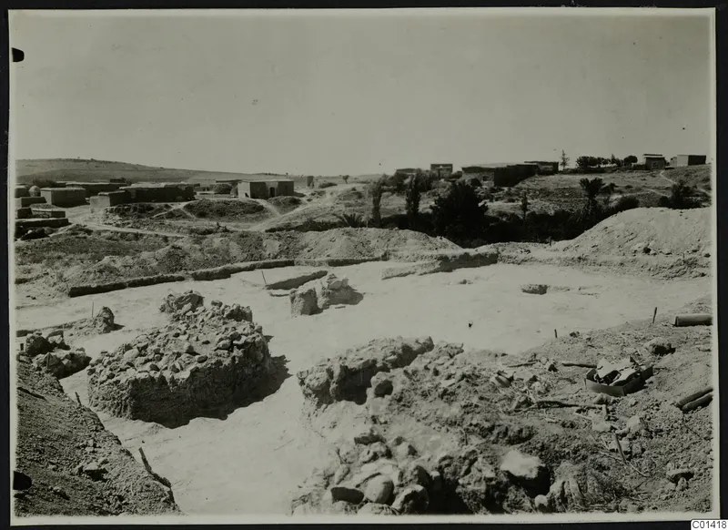 Excavated area with exposed stone and mud-brick walls at Ayios Iakovos, with a village of buildings on the hill behind