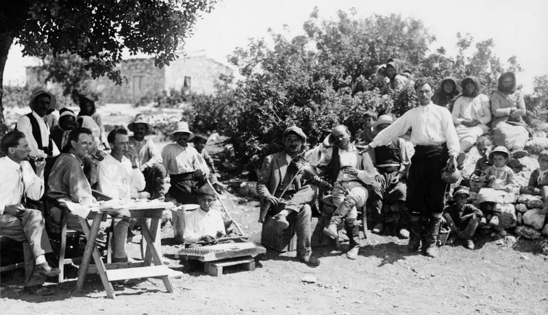 Outdoor party under a tree at Vouni, with a man playing violin and several men smoking pipes at a wooden table