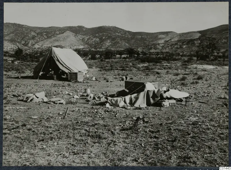 Tent, wooden boxes and draped bedding on bare ground at Ayios Iakovos, with a range of hills behind.