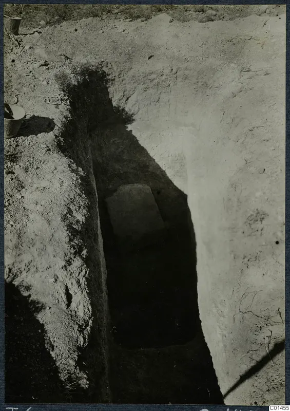 Tomb 6 at Ayios Iakovos during excavation, with a flat stone slab in a deep narrow cut and a bucket at the edge