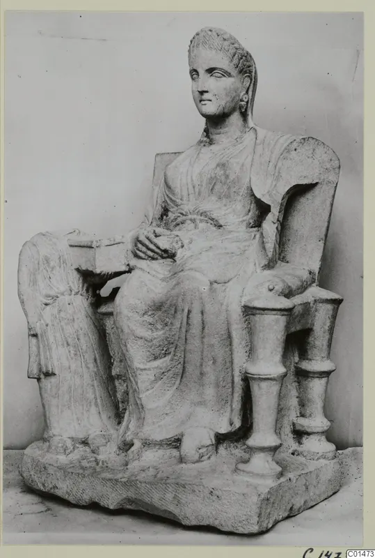 Seated female statue in stone on a throne with turned armrests, shown from the front left.